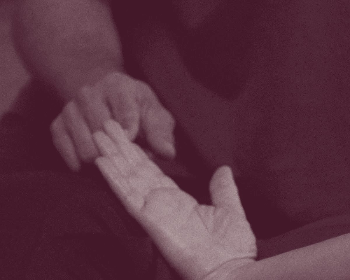 Two hands in warm light against a dark background, one palm open, the other reaching towards it
